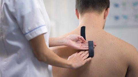 Doctor applying Y-shaped tapes on patient upper back, muscle spasm reducing