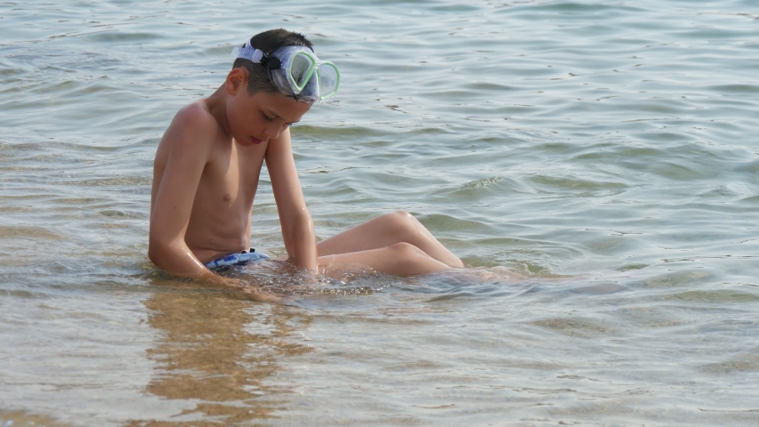  Exciting view of a happy boy in a mask sitting and entertaining in sea waves on a Turkish resort in summer in slow motion