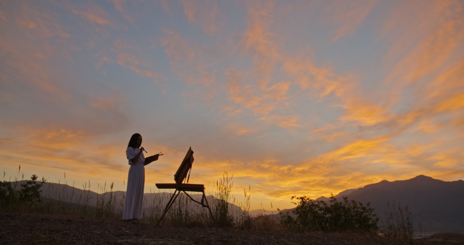Professional female artist is inspierd by beautiful mountain lake in sunset, creating a painting on easel, enjoying her hobby - recreational pursuit, inspirational landscape concept 4k footage