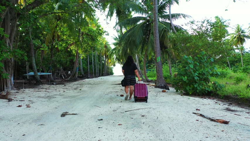 A woman pulling a purple trolley bag while walking in the middle of a sandy way under palm trees. On an island of the Maldives. Dolly in, wide angle, getting closer