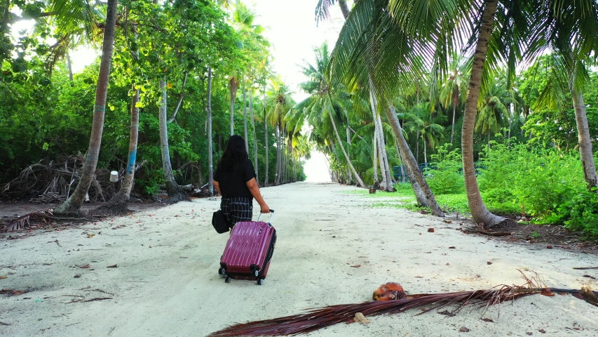 A woman pulling a purple trolley bag while walking in the middle of a sandy way under palm trees. On an island of the Maldives. Dolly in, wide angle.