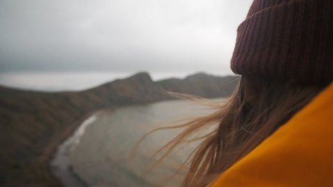 Back view of young woman hiker exploring nature in a dark misty rainy landscape. Mountains on the sea shore. Hiker girl wearing yellow raincoat trekking in cold stormy weather, hair blowing in wind. Stock-video