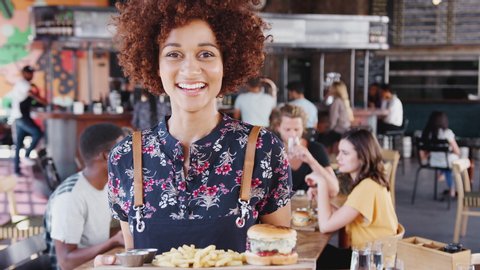 Portrait Of Waitress Serving Food In Busy Bar Restaurant Smiling At Camera