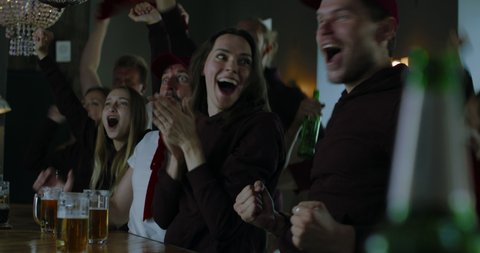 Group of male and female friends fans celebrating whilst watching game on TV screen in sports bar. 4K UHD RAW graded footage