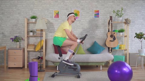 Funny tired athlete from the 80's with a mustache is engaged on a exercise bike in the house