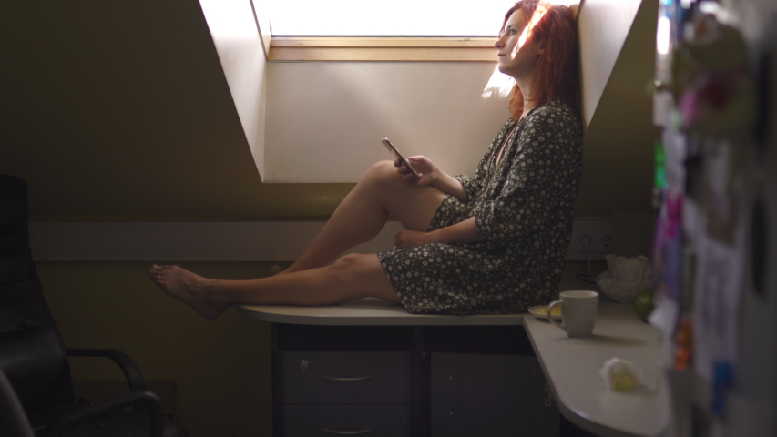 Texting: Positive smiling redhead woman using technology phone - Caucasian white girl wearing a dotted summer dress in an attic