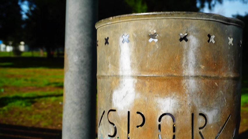 Metal garbage bin in a park engraved with the word SPQR with a view of a blurred park in the background