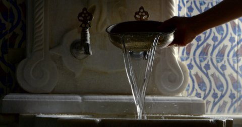 human is pouring water from bronze cup into stone washbasin in dark room, close-up