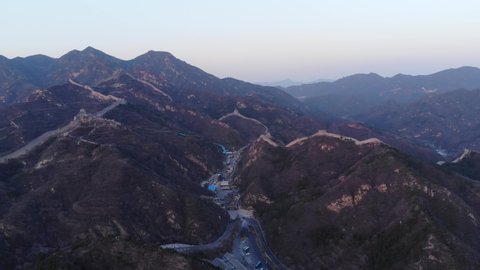Aerial view of Badaling Great Wall North in dusk, panoramic shot. Big mountains on background, tourist village lie at bottom of hills. Winding line of wall follow mountainous terrain