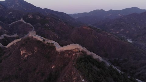 Aerial shot of Badaling Great Wall in evening twilight, stone strip lies on top of dark hills. Big mountains seen on background.