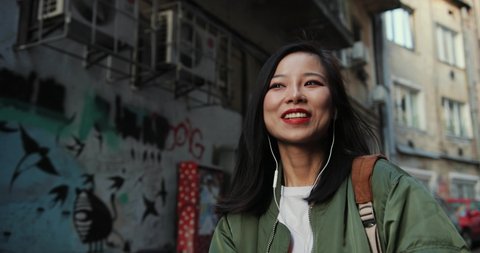 Asian happy and charming young stylish girl walking the street in slums and listening to the music in headphones with a smile.