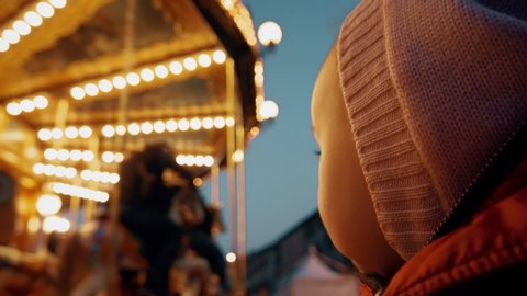 Amazed baby girl looking at illuminated retro carousel in the evening