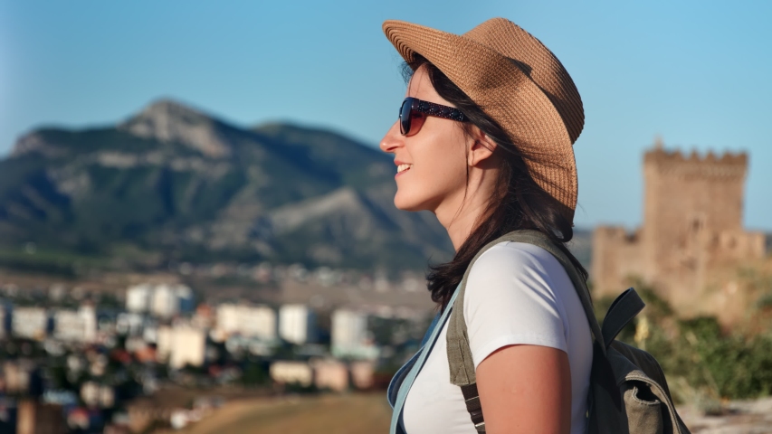 Smiling hipster young woman in hat and sunglasses admiring sunset at mountain landscape
