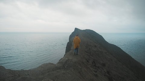 Back view of young woman hiker exploring nature in a dark misty rainy landscape. Mountains on the sea shore. Hiker girl wearing yellow raincoat trekking in cold stormy weather, hair blowing in wind. 库存视频