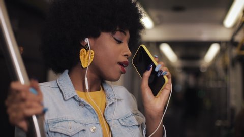 Portrait of young african american woman with headphones listening to music, sing and funny dancing in public transport. He holds the handrail.