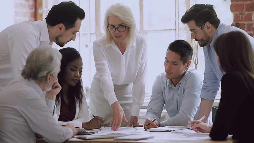 Diverse business team and old woman mentor leader brainstorm on paperwork talk engaged in teamwork at corporate briefing, senior boss explain new project plan training staff people at group meeting