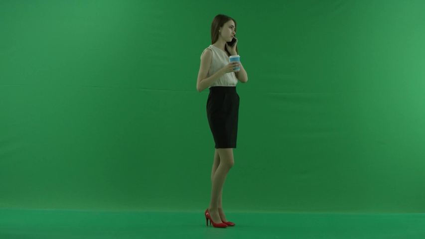 business woman is talking on her mobile cell phone with a cup of coffee or tea. She wears formal dress: white shirt and blak skirt. Businesswoman is standing over green screen