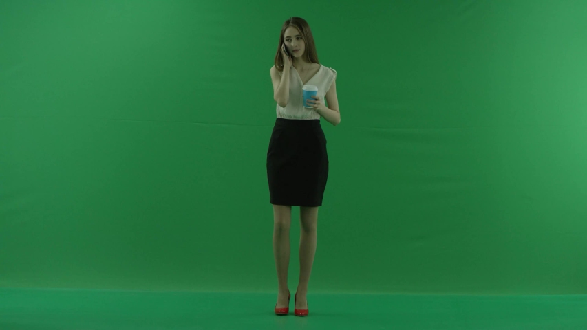 business woman is talking on her mobile cell phone with a cup of coffee or tea. She wears formal dress: white shirt and blak skirt. Businesswoman is standing front over green screen