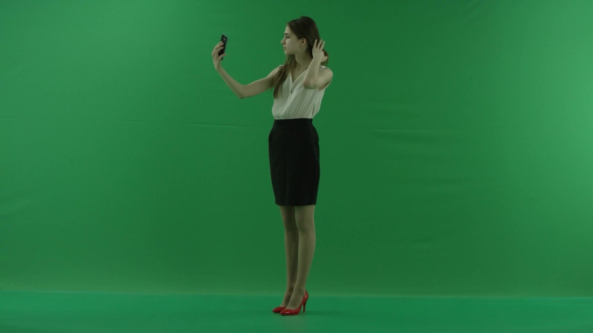 a happy pretty young business woman is making selfies. She wears formal dress: white shirt and blak skirt. Businesswoman standing over green screen