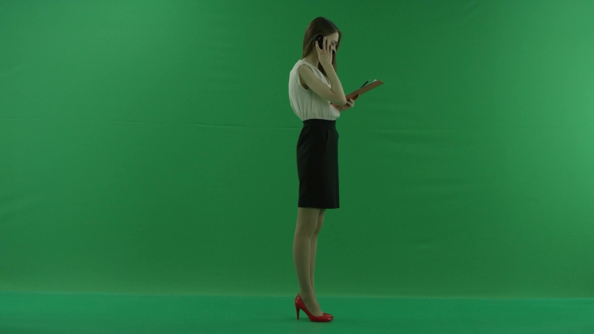 business woman is talking on her mobile cell phone and walking with folders. She wears formal dress: a white shirt and a blak skirt. Businesswoman is standing sideways over green screen