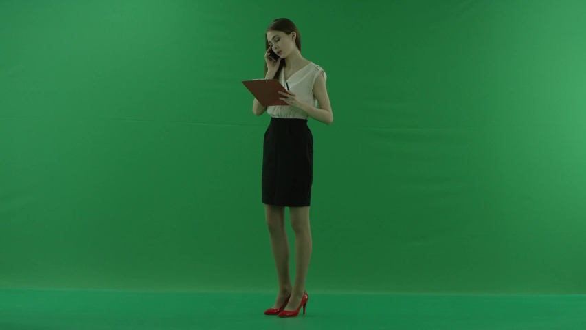 business woman is talking on her mobile cell phone and walking with folders. She wears formal dress: a white shirt and a blak skirt. Businesswoman is standing left side over green screen