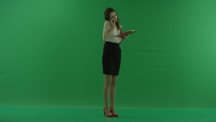 business woman is talking on her mobile cell phone and walking with folders. She wears formal dress: a white shirt and a blak skirt. Businesswoman is standing sideways over green screen