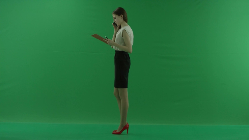 business woman is talking on her mobile cell phone and walking with folders. She wears formal dress: a white shirt and a blak skirt. Businesswoman is standing left side over green screen