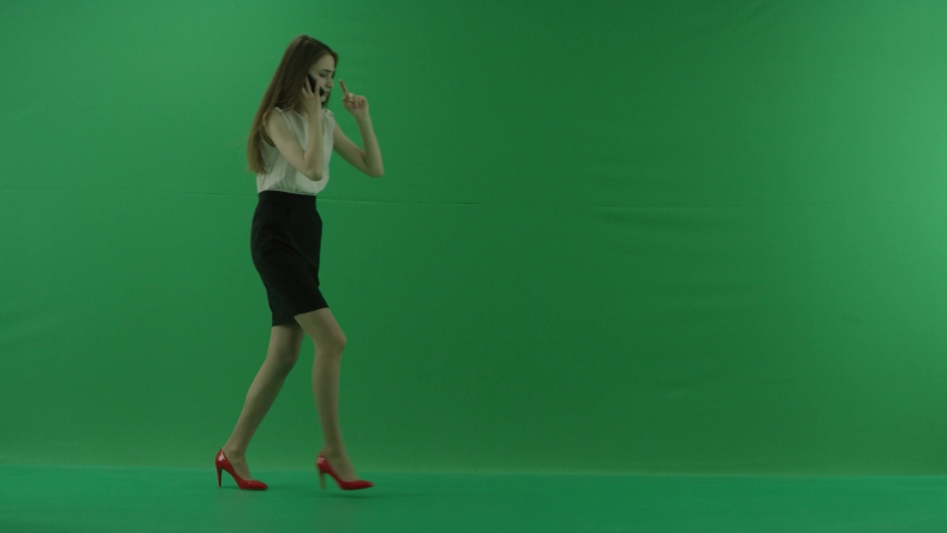 business woman is talking on her mobile phone and walking with a cup of coffee in the hand. She wears formal dress: white shirt and blak trousers. Businesswoman is over green screen