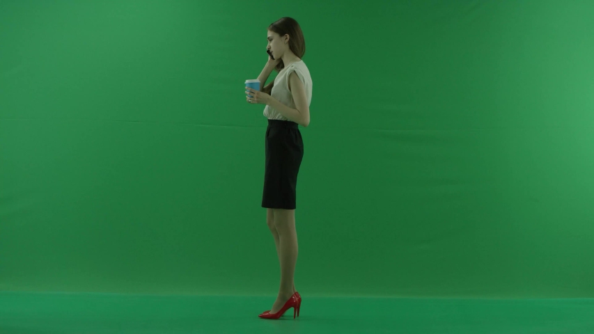 business woman is talking on her mobile cell phone with a cup of coffee or tea. She wears formal dress: white shirt and blak skirt. Businesswoman is standing over green screen