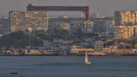 LISBON, PORTUGAL - APRIL 31, 2019: City scene with sailboat in the river and abandoned dock crane Lisnave dominating over the houses, view in warm sunset light