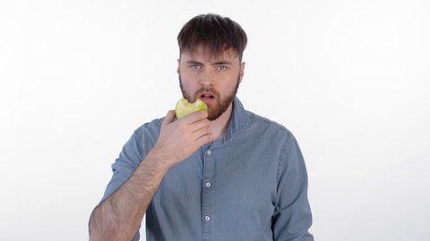 Medium shot of young bearded man standing isolated on white background, eating green apple and looking at camera