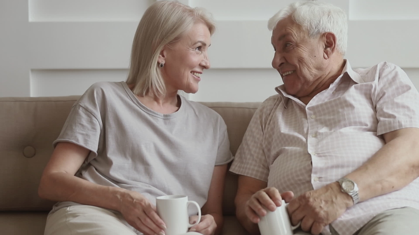 Happy old senior couple talking laughing drinking tea sit on sofa together, positive elder mature family man and woman bonding having fun spending time chatting joking relaxing on couch at home | Shutterstock HD Video #1032303668
