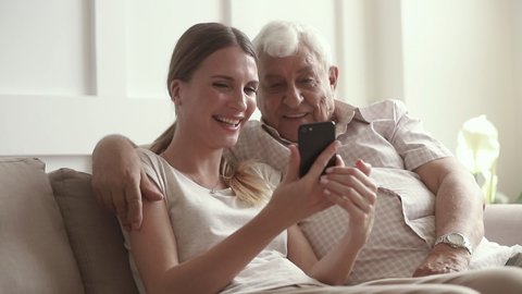 Happy family senior retired father embracing adult daughter using smartphone funny apps at home, cheerful old elder dad hug young grown woman talking laughing holding phone having fun sit on sofa