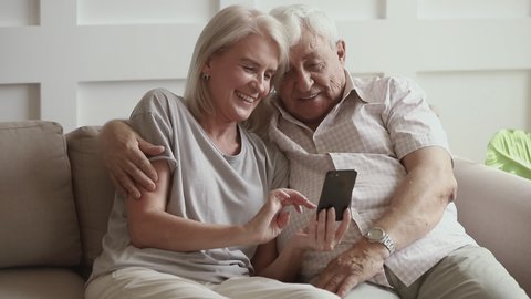 Happy old senior couple laughing using smartphone funny app looking at screen, smiling retired husband and middle aged wife talking having fun watching photos on mobile phone on sofa at home
