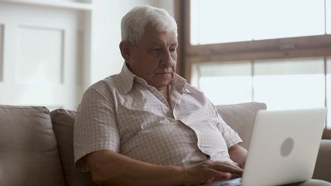 Senior older mature man typing on laptop laughing browsing internet using apps sit on sofa alone, elder aged retiree working on computer looking at screen at home, retirement and technology concept