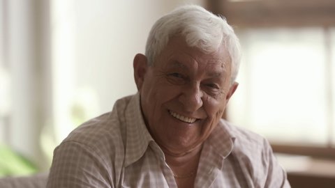 Senior old gray-haired man looking at camera, serious happy elder mature grandfather with toothy smile posing for close up video portrait alone at home or retirement house, older people health care