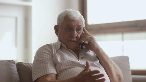 Happy elder senior man holding cellphone talking on phone at home, smiling older retired mature grandfather making call enjoy mobile telecommunication laughing speaking by smartphone sit on sofa