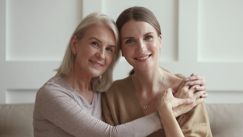 Smiling mature middle aged loving mum embracing grown young adult daughter bonding looking at camera, happy two generation women family laughing hugging together sit on sofa at home, portrait Royalty-Free Stock Footage #1032303752