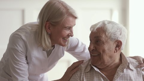Smiling mature kind woman nurse doctor talking to happy disabled old elder man, happy female therapist cheering taking care of senior patient giving support and medical assistance in retirement house