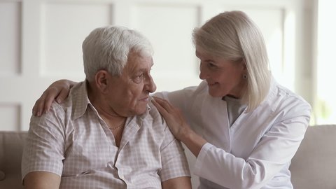 Kind mature woman nurse caregiver supporting talking to old elder man help with problem, female doctor therapist in uniform on sofa at home hospital cheering give empathy listen to senior patient
