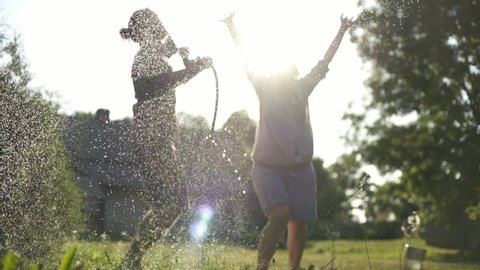 Young cheerful beautiful couple hosing the garden and playing under sprays of water at the sunset time. Summer happiness and love concept