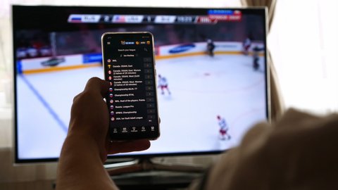 Riga, Latvia - 06/06/2019 EDITORIAL: Against the background of a TV, a man watches a sports channel and places bets using a mobile application. Concert of: At Home, At the Bar, Football, Sport.
