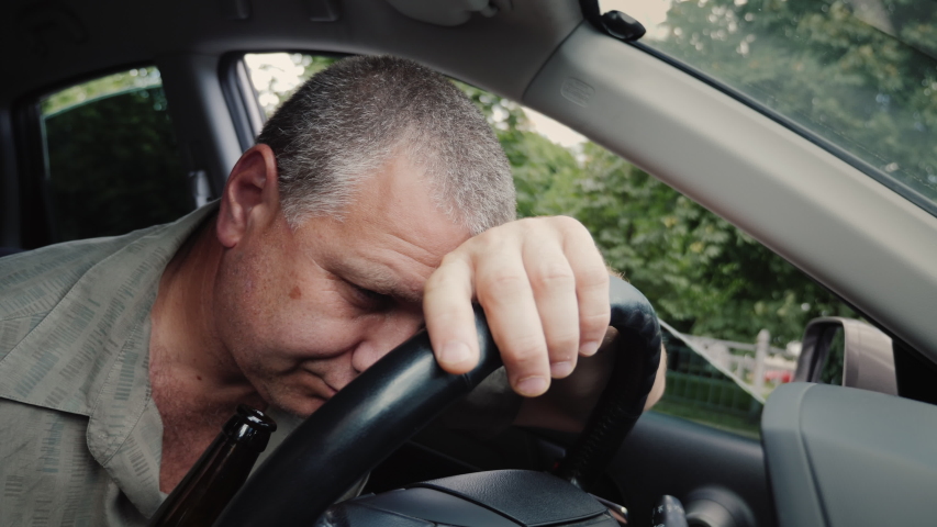Drunk driver with a bottle of alcohol in his hand dozing on the steering wheel