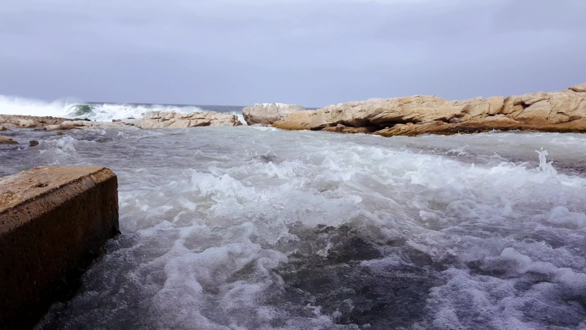 Flowing foam water running along channel towards ocean waves under stormy cloudy sky. Low angle perspective.