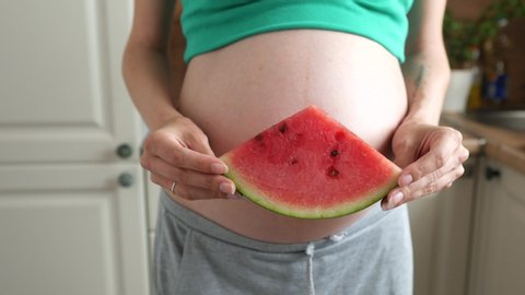 close-up of a pregnant woman holding and biting a piece of watermelon standing in the kitchen