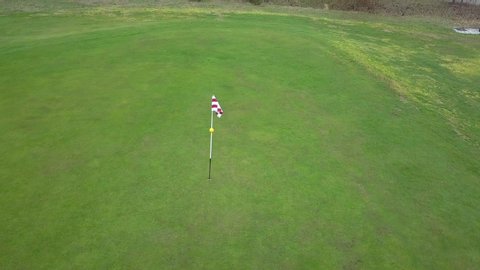 Panoramic drone shot of a red and white golf flag,ing in the wind, on a bright green golf field, on a sunny day