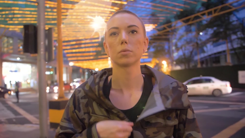 Woman with shaved head standing on street pulls hood of camouflage jacket over her head. Slow motion view of lady pulling camo jacket up to cover her head.