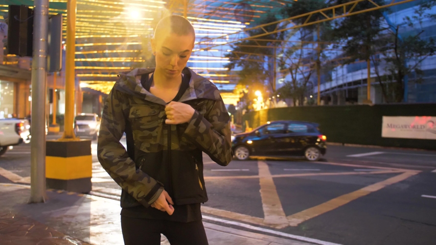 Attractive, thin woman standing outside by the street puts on her camouflage jacket. Lady wearing camo rain jacket zips up while waiting for a taxi.