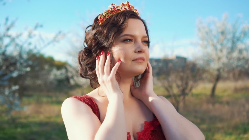 portrait of adult girl with dark hair and bright blue eyes, young attractive woman with gentle natural make-up and red nails tilts head to side, queen poses in chic dress in front of clear azure sky