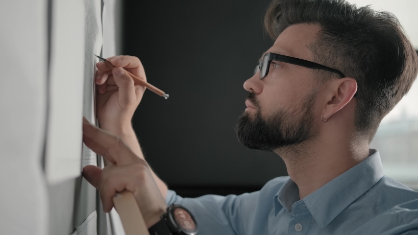 Bearded man with glasses makes notes on blueprints on the loft-style wall. Stylish office in loft style: brick wall with notes and blueprints. Designer works in loft office and makes notes. Close up Royalty-Free Stock Footage #1032347579